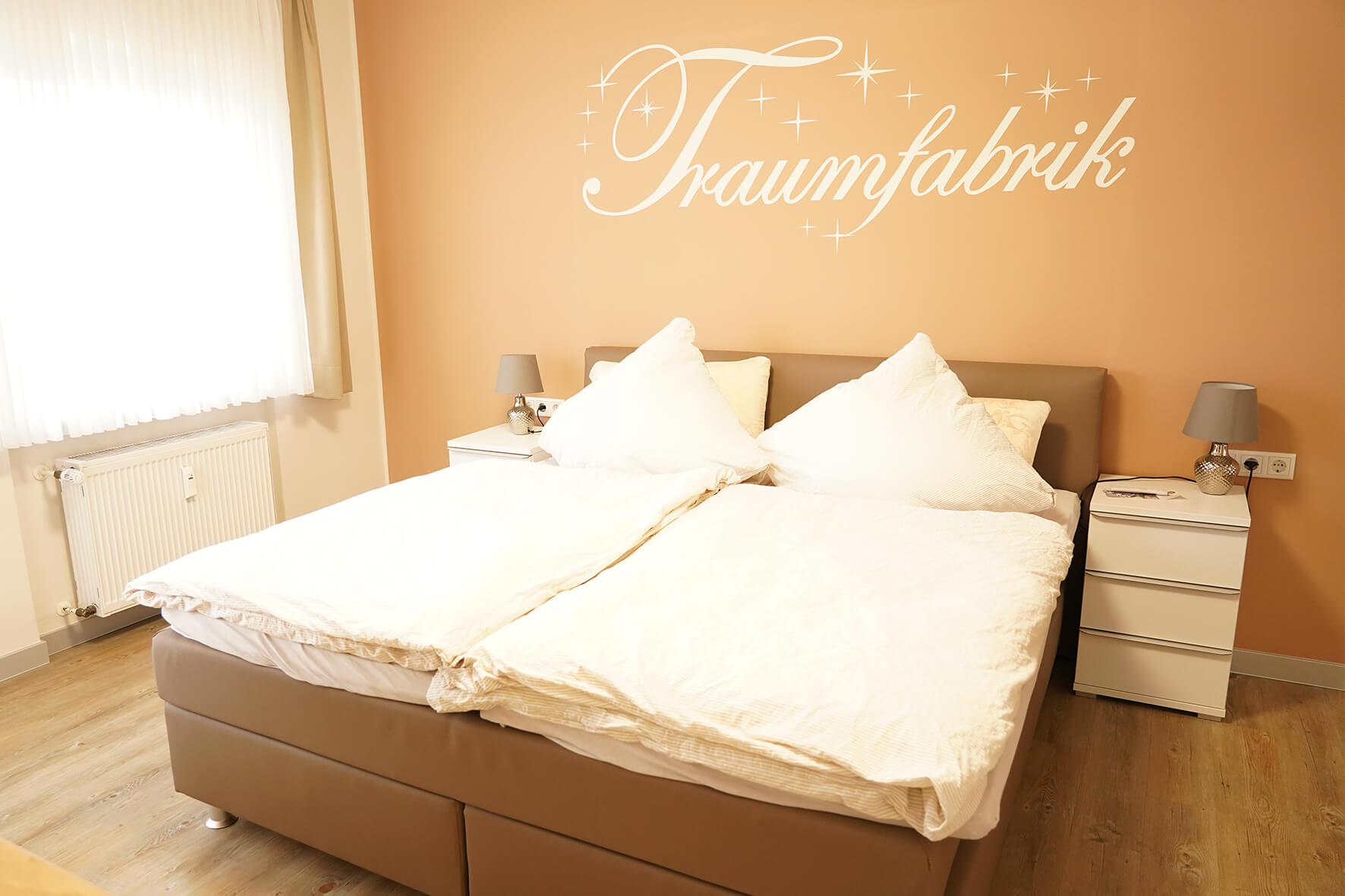 Zimmer mit einer Pfirsichfarbenden Wand und einem Wandtattoo "Traumfabrik" mit einem braunen Lederbett davor