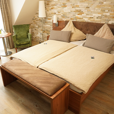 Braunes Holz Bett mit beiger Bettwäsche vor einer Stein Wand. In der Ecke steht ein grüner Samtsessel.